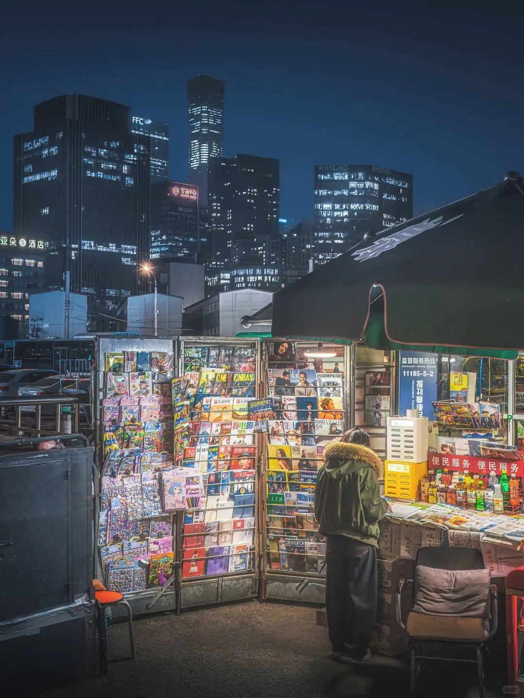 A nighttime cityscape with illuminated buildings and a street vendor stall selling various items.