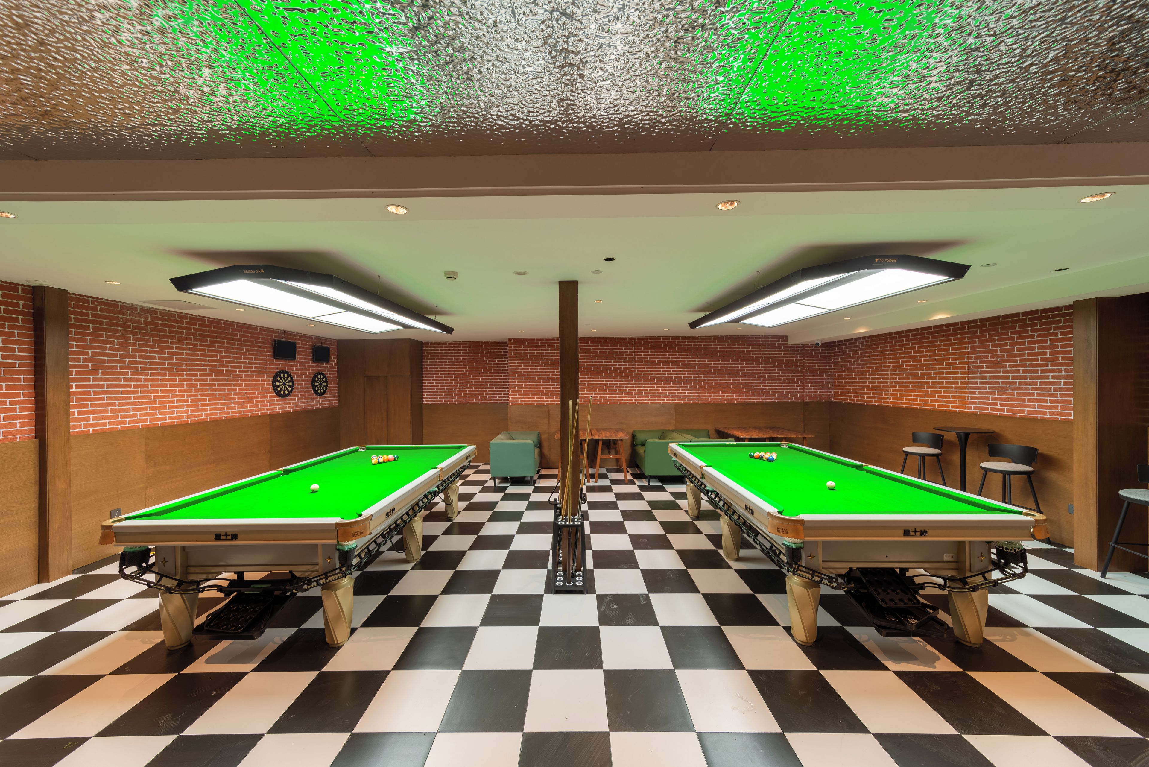 A room with two pool tables, checkered flooring, brick walls, and ceiling lights.