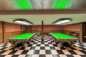 Una sala con dos mesas de billar, piso a cuadros, paredes de ladrillo y luces en el techo.