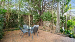 An outdoor patio with a glass table and chairs, surrounded by trees and a stone wall. - Yowie Bay Waters | Pool, Spa & Port Hacking Views (Yowie Bay)