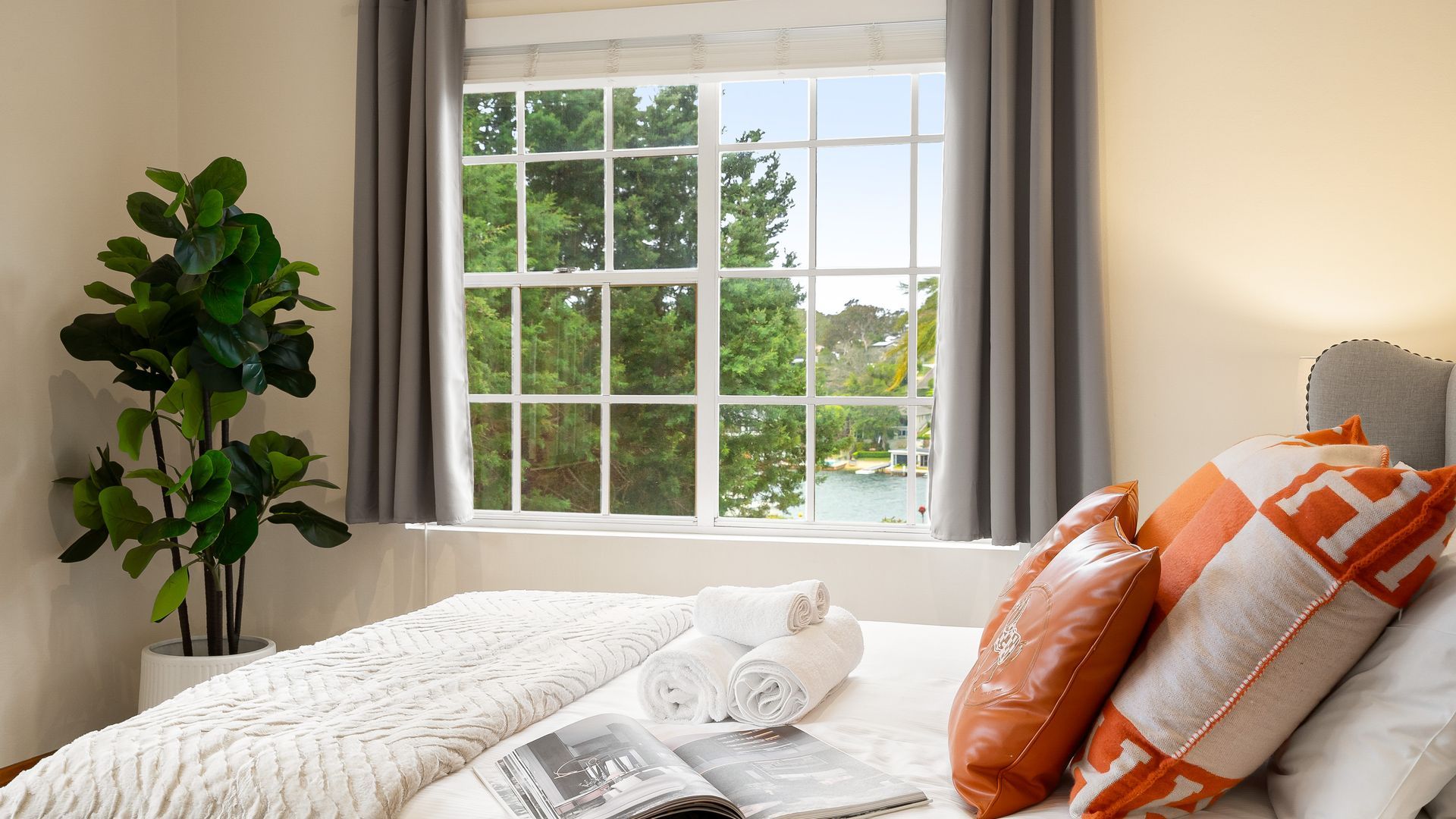 A neatly made bed with rolled towels, a magazine, and a view of a lake through a window with curtains.