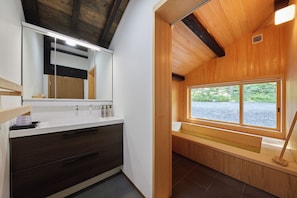 A modern bathroom with a wooden ceiling, a large mirror, a white sink, and a window overlooking a gravel area.