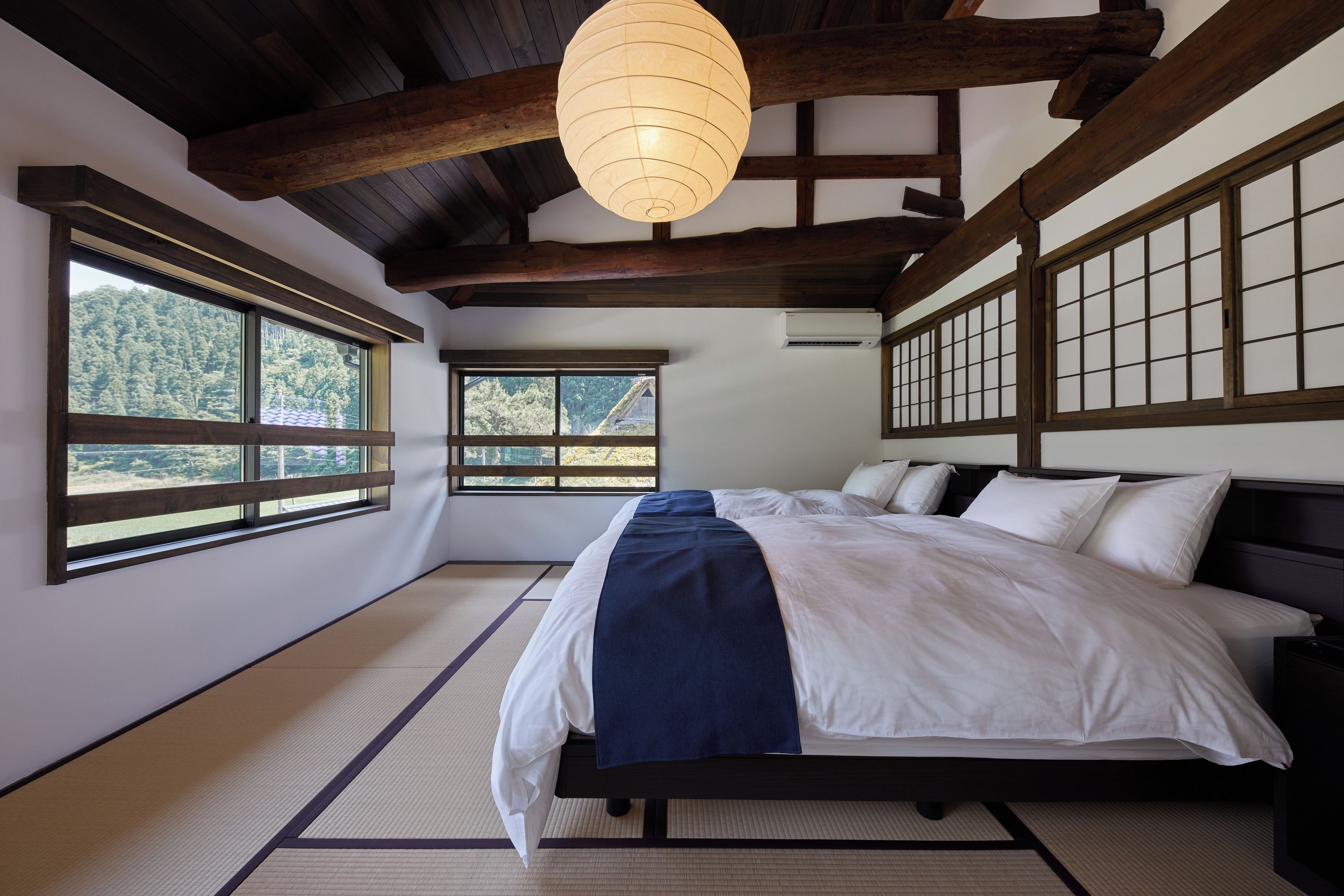 A traditional Japanese-style bedroom with tatami flooring, a bed with white and blue bedding, and large windows offering a view of the forest.