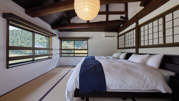 A traditional Japanese-style bedroom with tatami flooring, a bed with white and blue bedding, and large windows offering a view of the forest.