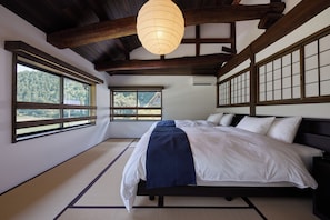 A traditional Japanese-style bedroom with tatami flooring, a bed with white and blue bedding, and large windows offering a view of the forest.