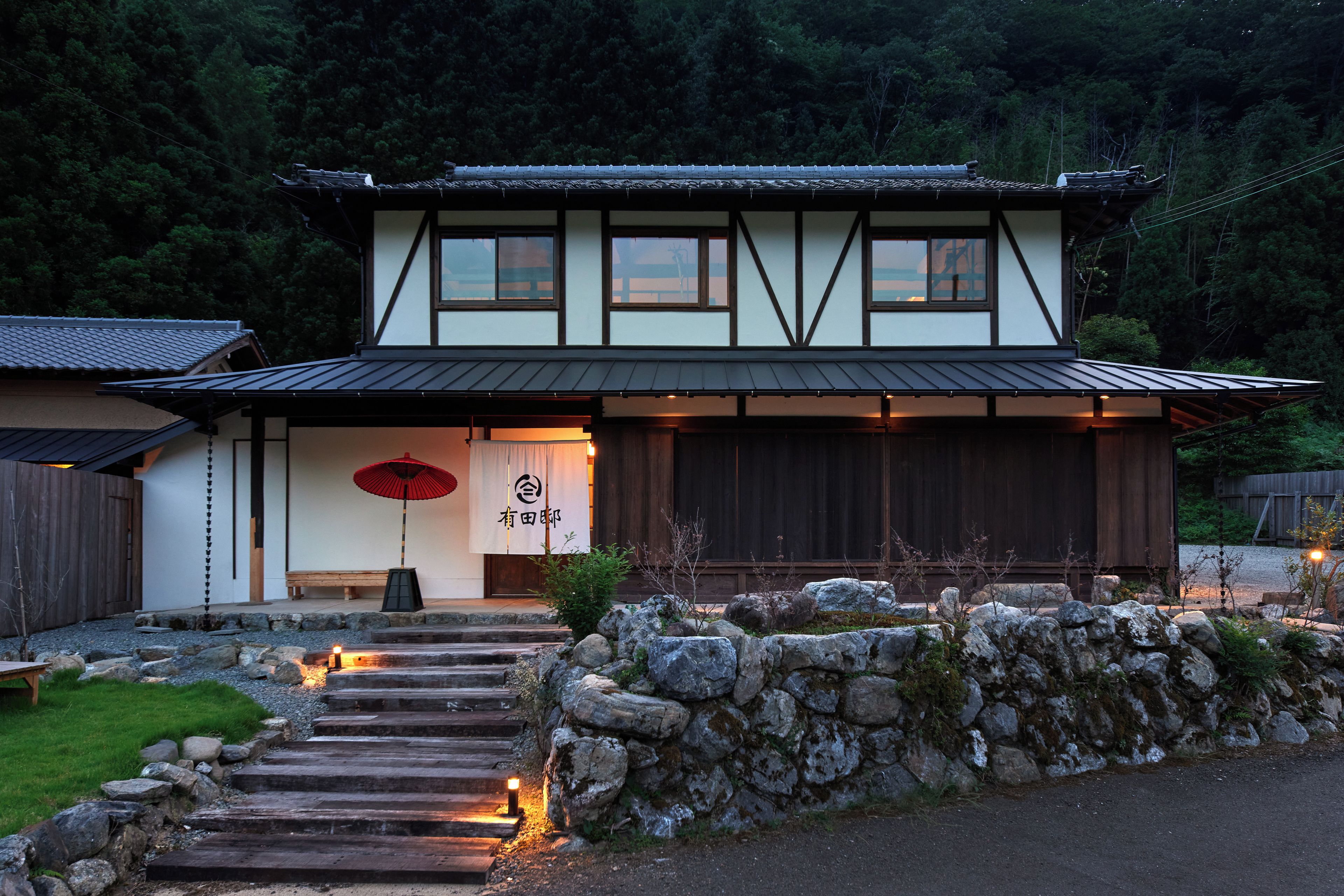A traditional Japanese building with a wooden exterior, a stone pathway leading to the entrance, and a red umbrella.