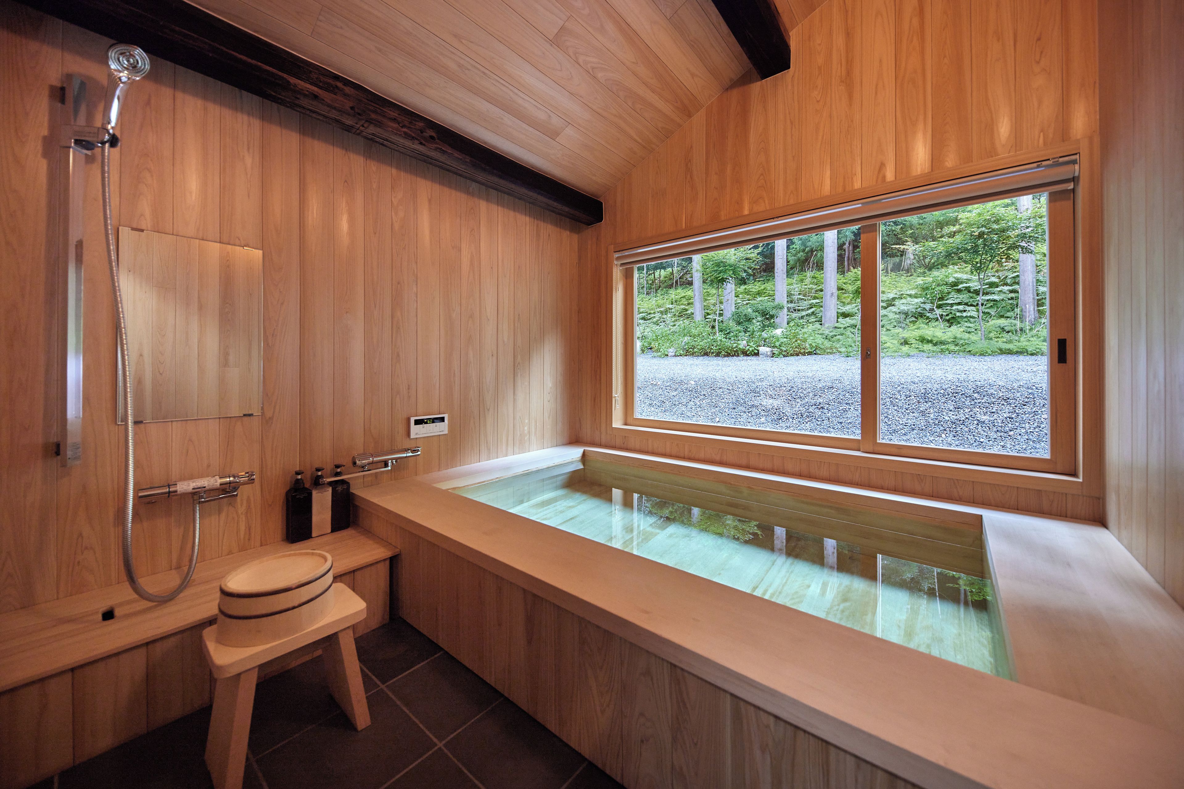 A wooden bathroom with a large bathtub, a shower, and a window offering a view of a forest.