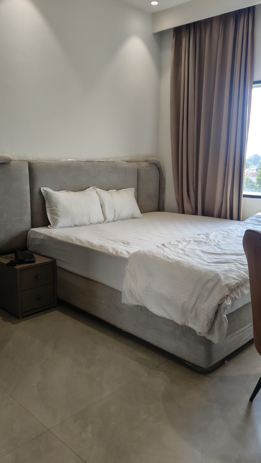 Habitación con una cama individual, cabecera gris, ropa de cama blanca, una mesita de noche con teléfono y una ventana con cortinas.