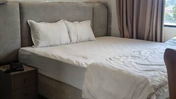 Habitación con una cama individual, cabecera gris, ropa de cama blanca, una mesita de noche con teléfono y una ventana con cortinas.