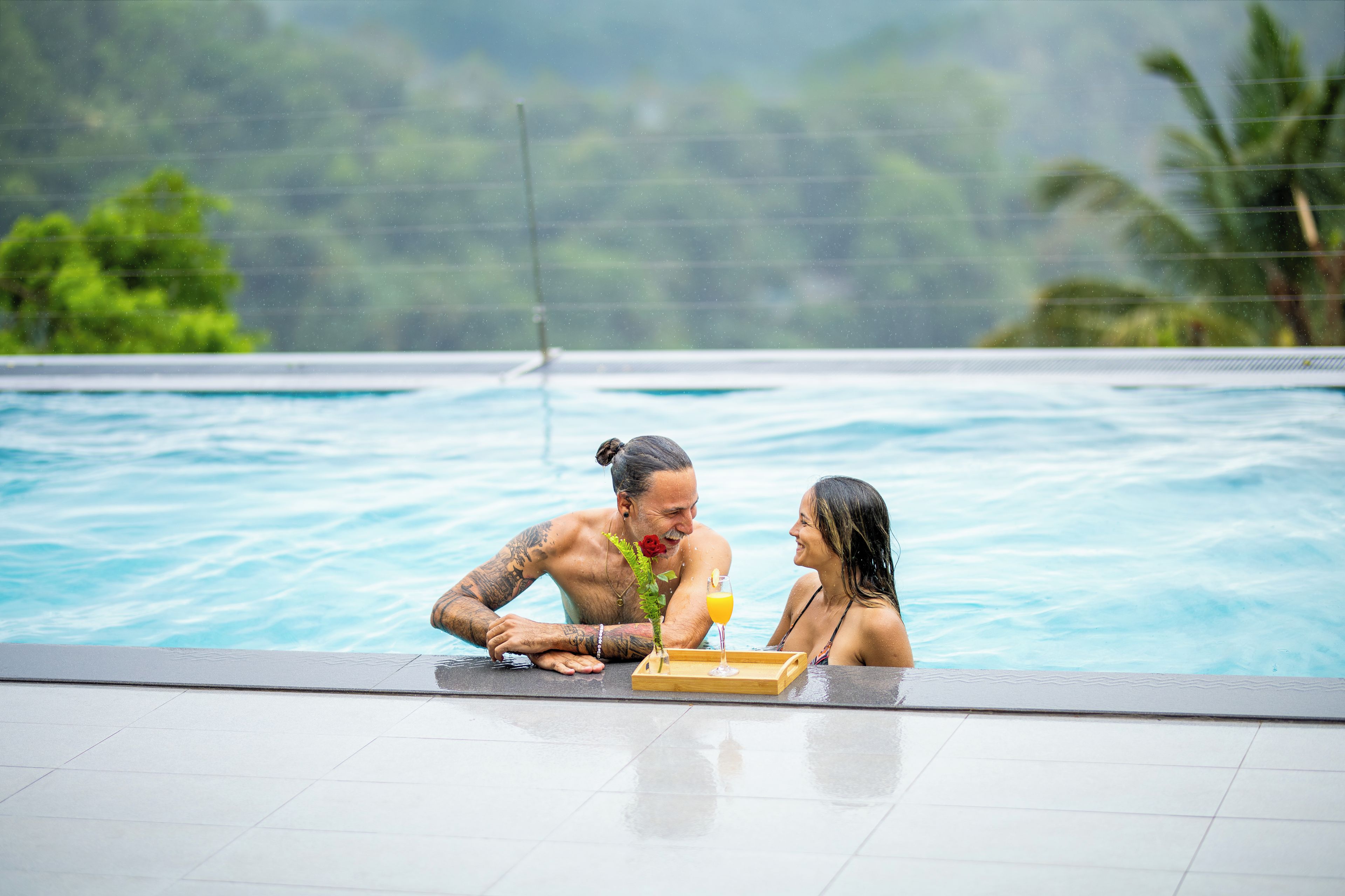 A couple in a swimming pool with a wooden tray and a drink.