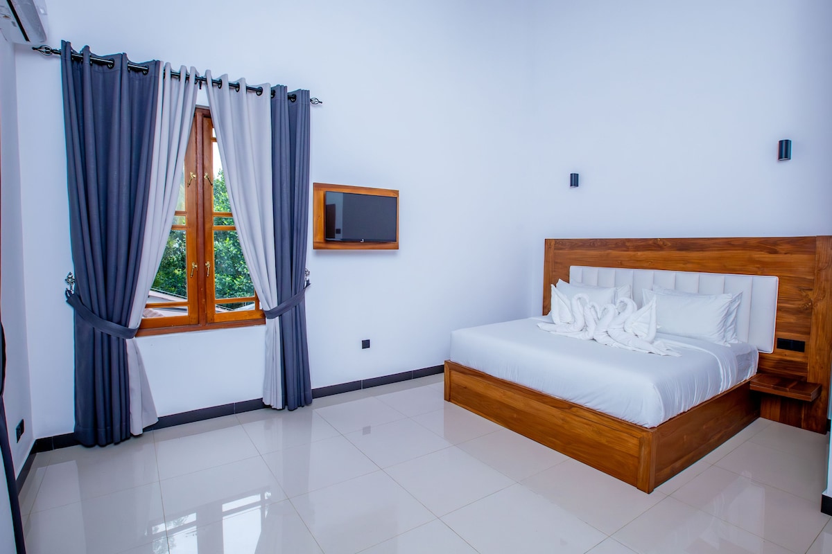 Un dormitorio con una cama de madera, ropa de cama blanca, una ventana con cortinas, un televisor de pantalla plana y suelo de baldosas.