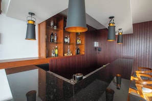 Zona de bar con mostrador de madera, estantes con botellas de licor e iluminación colgante.