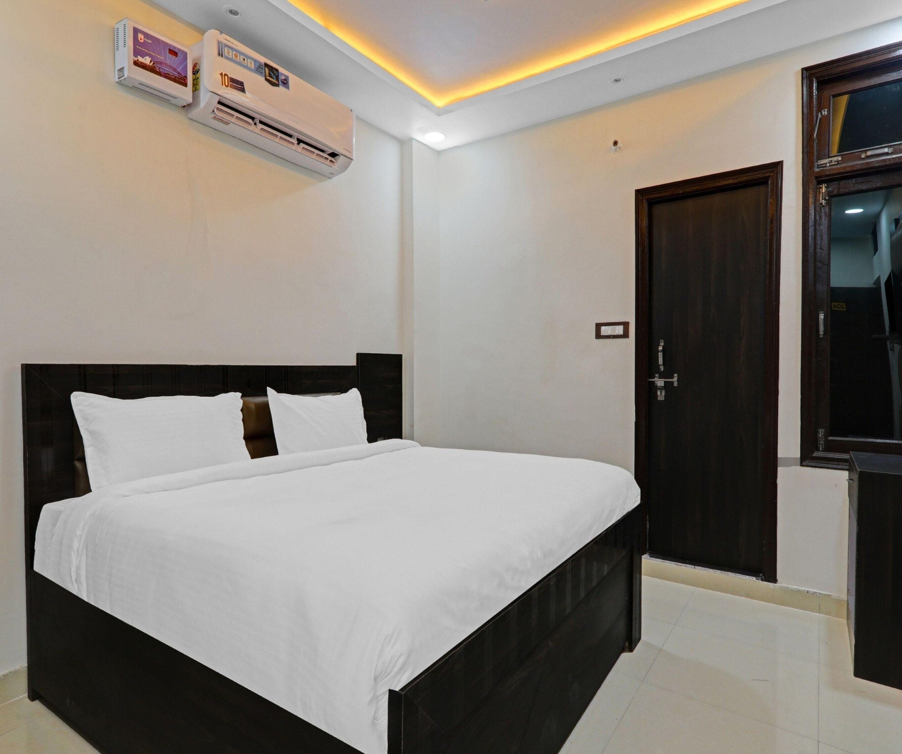 A bedroom with a bed, a dark wooden headboard, white bedding, an air conditioning unit, a door, and a light fixture on the ceiling.