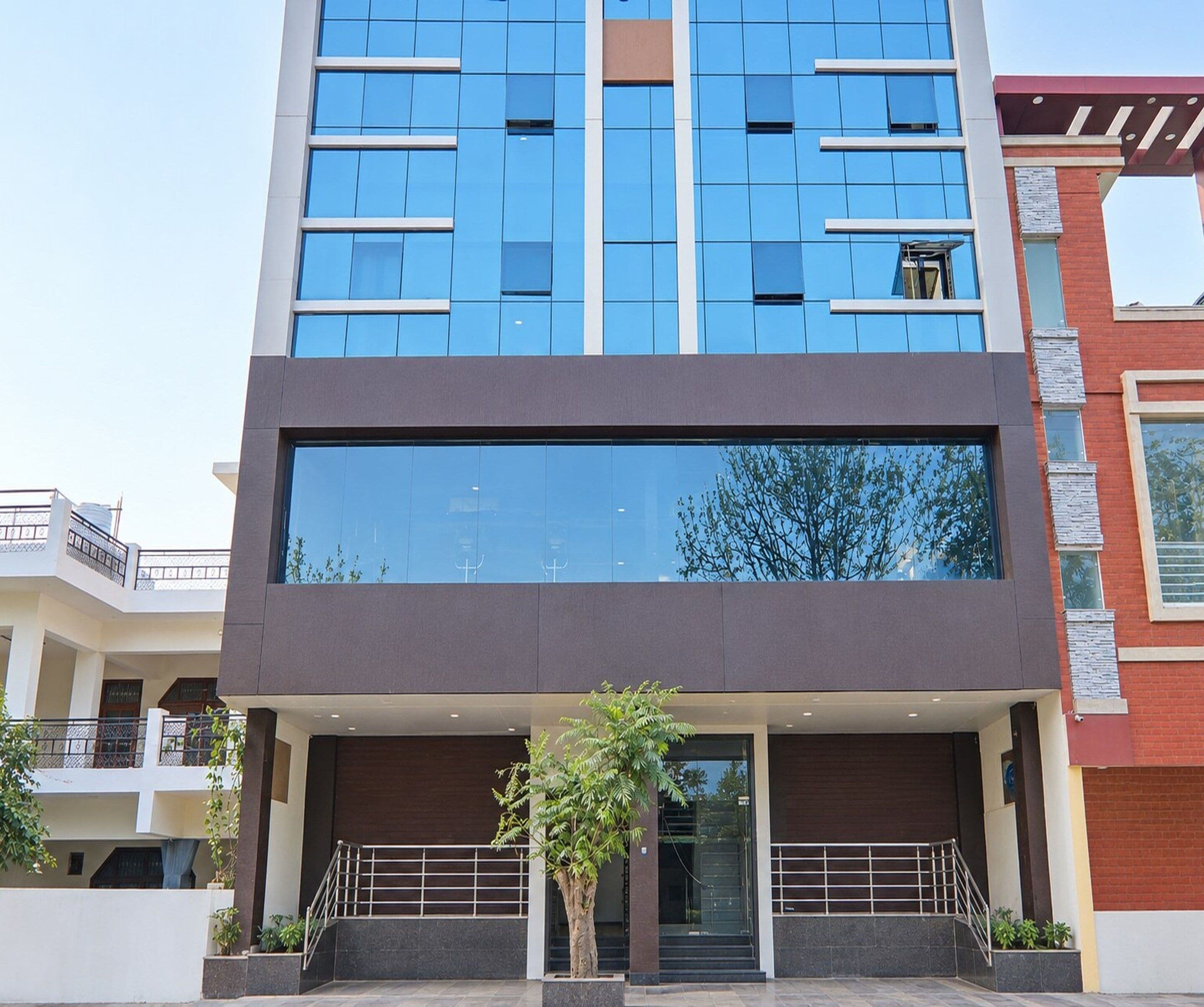 Un bâtiment moderne doté d’une grande façade en verre, d’une entrée centrale avec un escalier et d’un petit arbre au premier plan.
