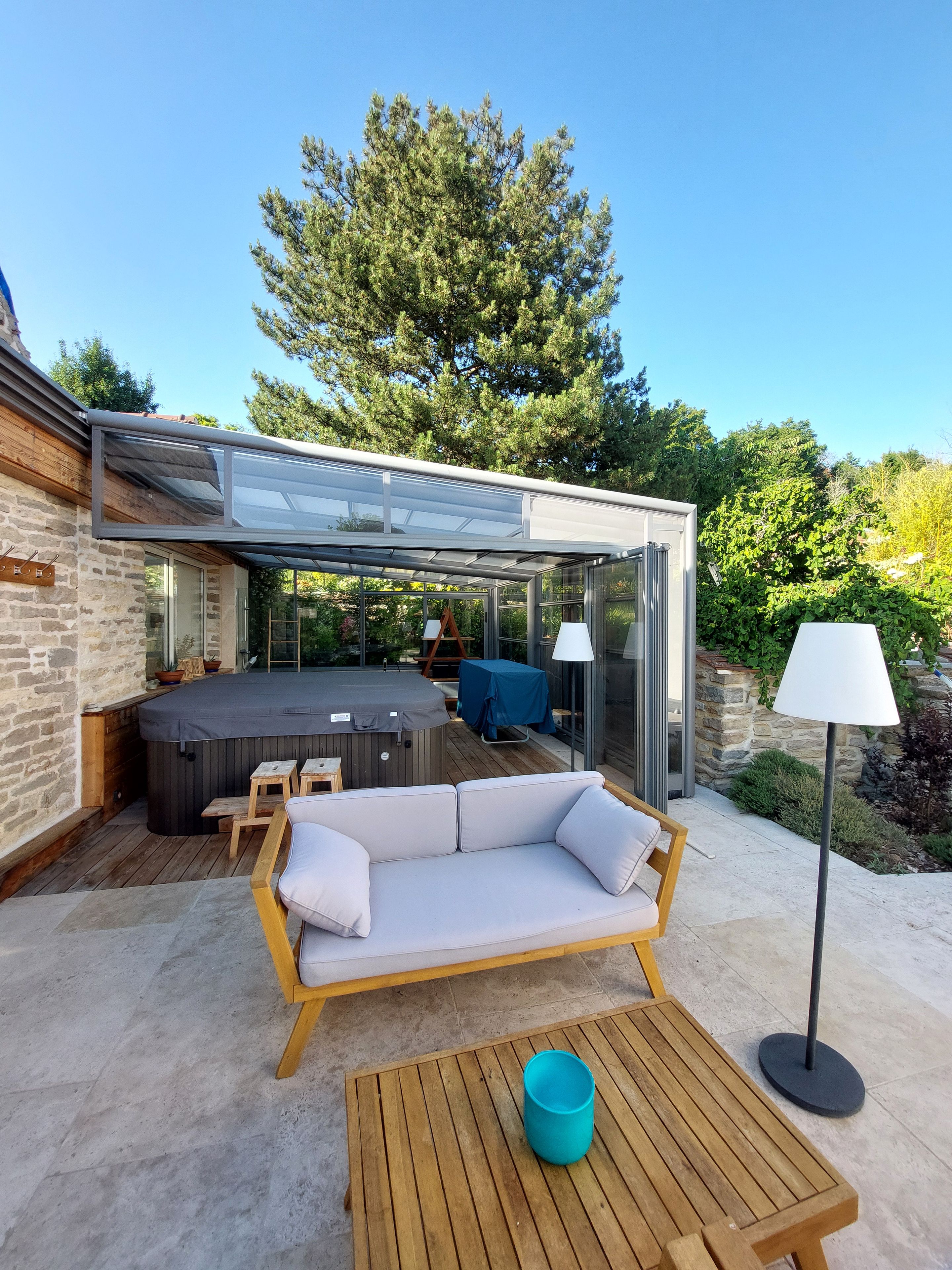 A modern outdoor patio with a glass-enclosed hot tub, a wooden sofa, and a table.