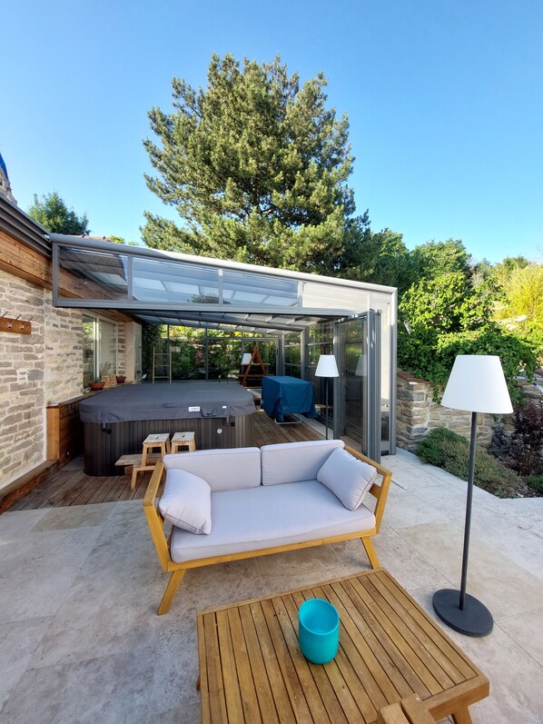 A modern outdoor patio with a glass-enclosed hot tub, a wooden sofa, and a table.