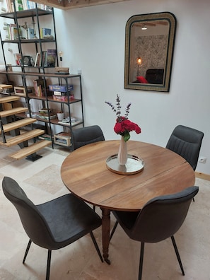 Une table de salle à manger ronde en bois, sur laquelle est posé un vase blanc rempli de fleurs, est entourée de quatre chaises noires. On aperçoit, au fond, une bibliothèque contenant divers objets.