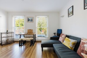 Una sala de estar moderna con piso de madera, un sofá, una mesa y una silla. Hay ventanas con persianas, un cuadro en la pared y una fotografía enmarcada sobre el sofá.