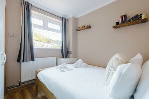Habitación de hotel con cama, ventana con cortinas, radiador, estante con libros y un interruptor de luz en la pared.