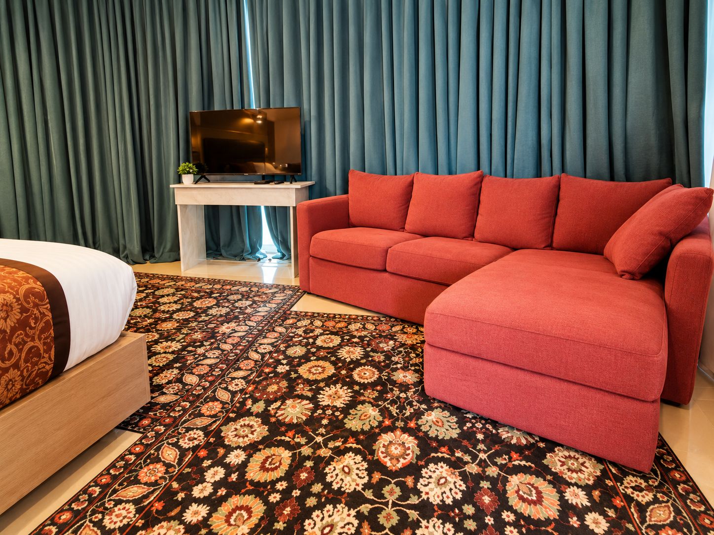 Una habitación de hotel con una cama, un sofá rojo, un televisor sobre un mueble bajo y una alfombra estampada.