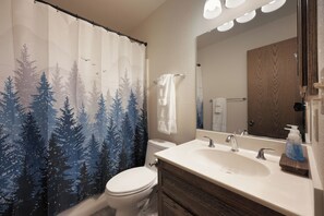 Ein Badezimmer mit einer Duschvorhang mit Waldmotiv, einer weißen Toilette, einem Waschbecken mit Spiegel und einer hölzernen Kommode.
