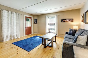A living room with a blue rug, a wooden floor, a grey sofa, a small table, and a door with a red frame. - Adirondack Cabin at Historic Fenton House Hotel (Lowville)
