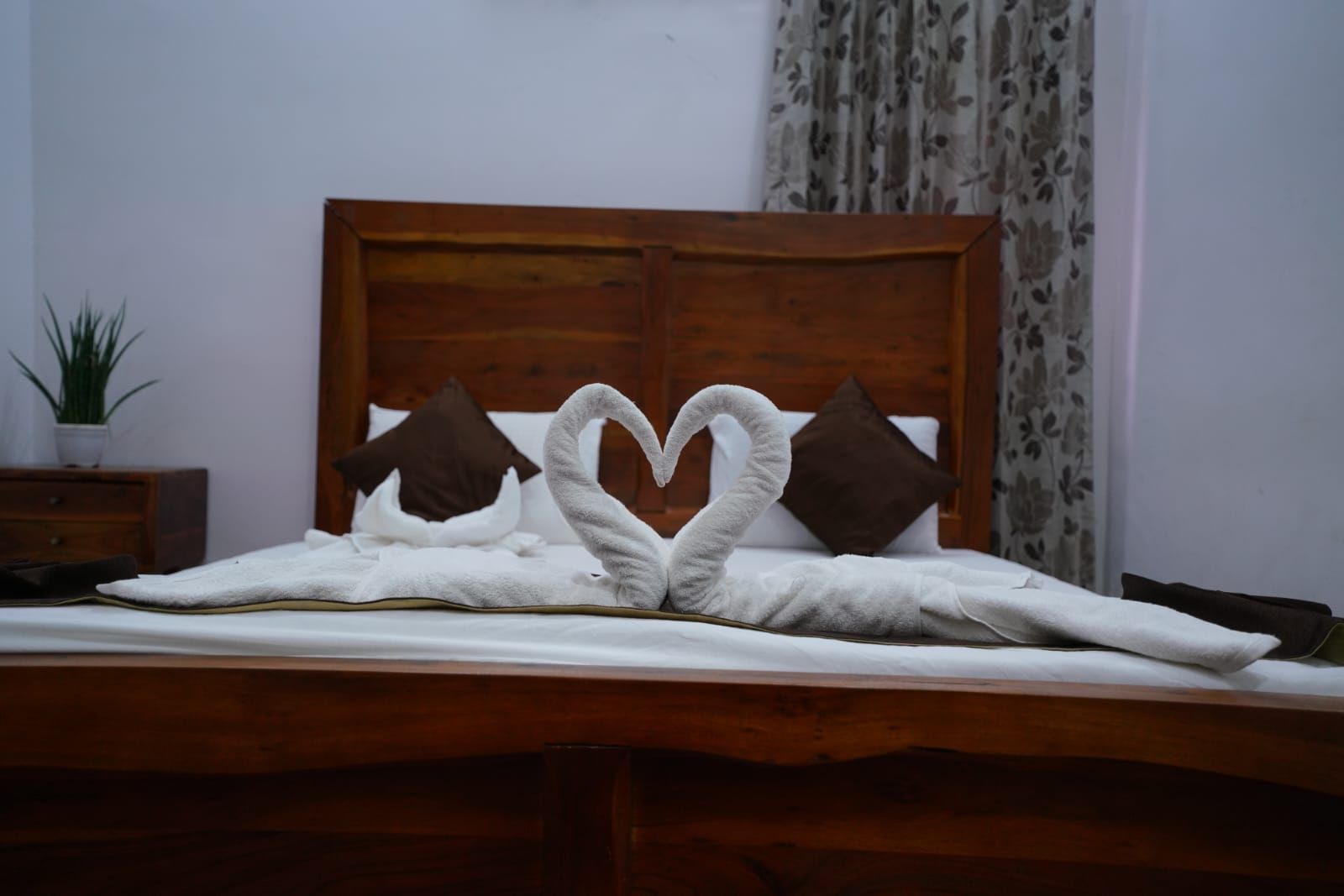 A bed with a wooden headboard, two pillows, and a towel arranged in a heart shape.