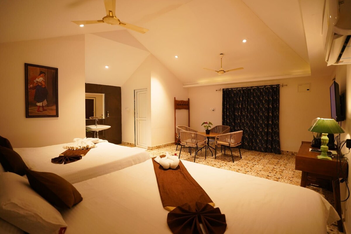 A hotel room with two beds, a dining table with chairs, a ceiling fan, and a television.