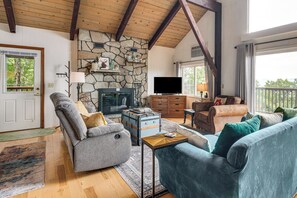 Ein gemütliches Wohnzimmer mit Kamin aus Stein, Holzbalken, Fernseher und bequemen Sitzmöglichkeiten.