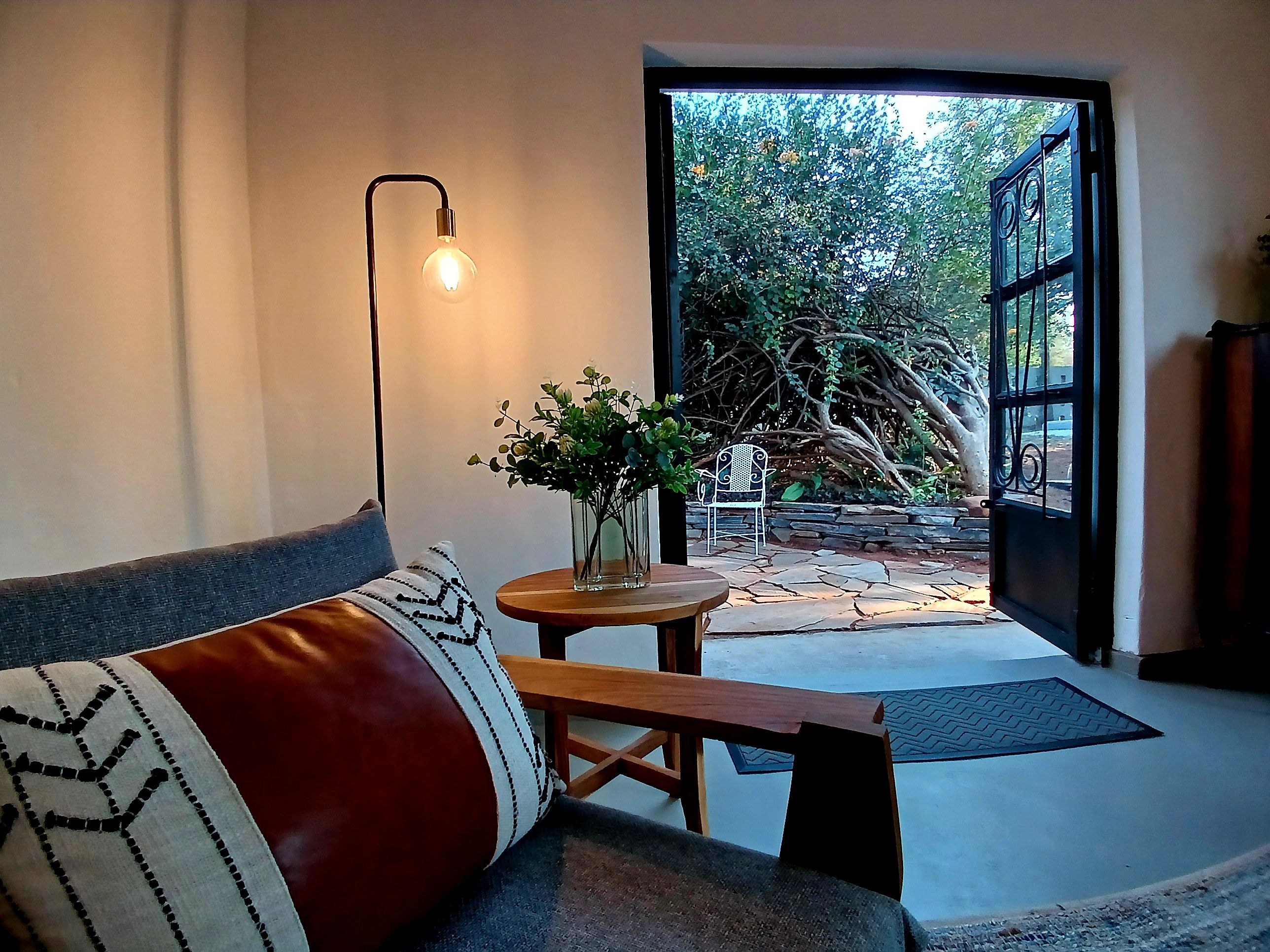 A modern interior with a sofa, a table with a vase of flowers, and a view of a garden through an open door.