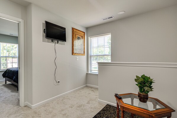 A room with a bed, a television mounted on the wall, a framed picture, a window with blinds, and a small table with a plant.