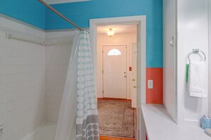 A bathroom with white tiled walls, a shower curtain, a door leading to a hallway, and a towel rack with a towel. - 2 Mi to Dtwn Pensacola: Historic Home w/ Patio! (Pensacola)