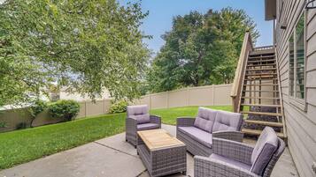 Une terrasse dotée de meubles en osier et d’une table en bois, entourée d’une pelouse bien entretenue et d’arbres.