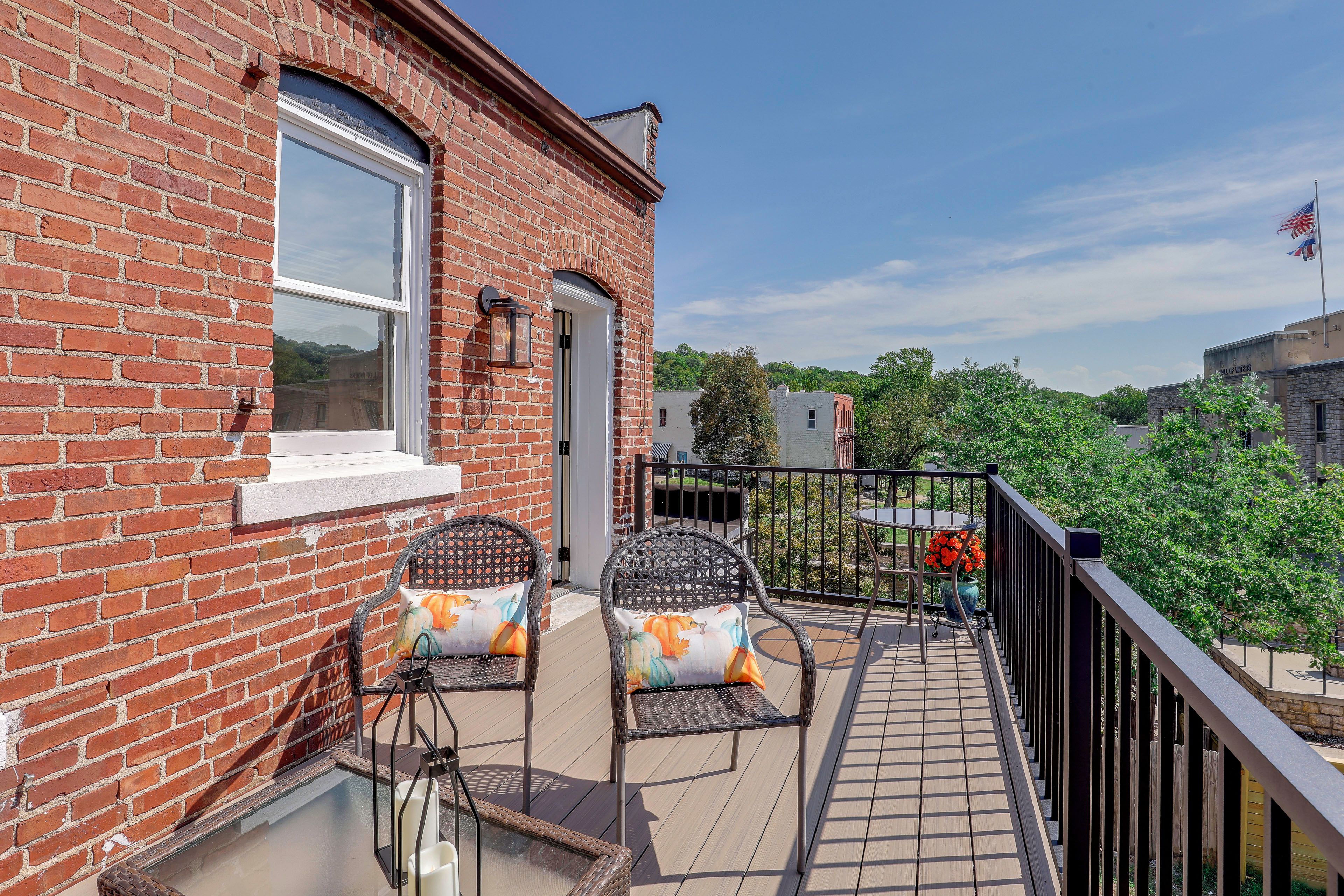 A brick building with a balcony featuring two wicker chairs with cushions, a small table, and a railing.