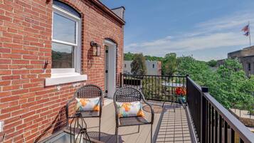 A brick building with a balcony featuring two wicker chairs with cushions, a small table, and a railing.