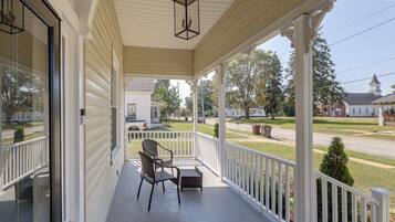 A covered porch with a chair and a small table, overlooking a street with houses and a church.