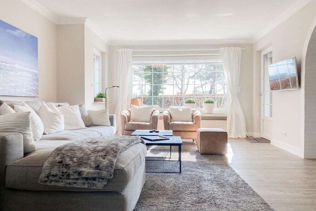 Una sala moderna con un sofá, sillones y una mesa de centro. Hay un ventanal con cortinas, un cuadro en la pared y vistas a árboles.