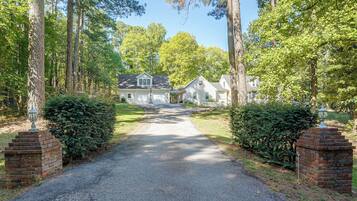 A gravel driveway leading to a residential area with houses, surrounded by trees and a white picket fence.
