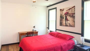A bedroom with a bed, a desk, a chair, a ceiling fan, and a window with blinds.