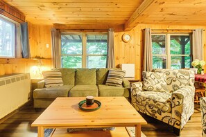 A cozy cabin interior with a sofa, patterned armchair, and a table with a mug. - Waterfront Maine Getaway: Deck + Fire Pit (Embden)