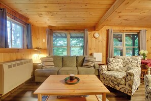 Interior acogedor de una cabaña con techo de madera, un sofá, un sillón estampado, una mesa con una taza de café y ventanas amplias con cortinas.