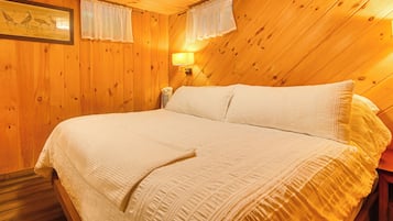 Una habitación con techo de madera inclinado, una cama con ropa blanca, una lámpara fijada a la pared y un cuadro enmarcado sobre la cama.