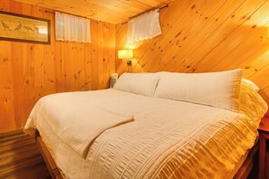 A room with a wooden sloped ceiling, a bed with white bedding, a wall-mounted lamp, and a framed picture above the bed.