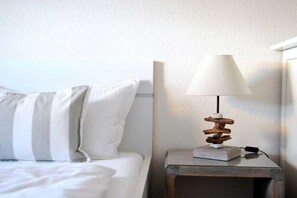 A bed with white bedding and pillows, a bedside table with a lamp and a book, and a wall with a textured finish. - Sonnenkuhle - Ameland 09 (Wenningstedt - Braderup)