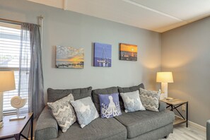 A living room with a grey sofa, decorative pillows, and three wall-mounted paintings depicting coastal scenes. - Shop, Dine, Walk to Beach! South Padre Island Unit (South Padre Island)