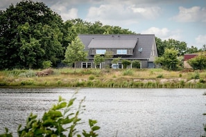 A house with a sloped roof, surrounded by trees and a body of water. - Holiday apartment with 1 bedroom (Aurich)