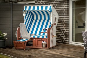 A beach chair with a striped cover, a small wooden table, and a trash bin on a wooden deck. - Holiday apartment with 1 bedroom (Aurich)