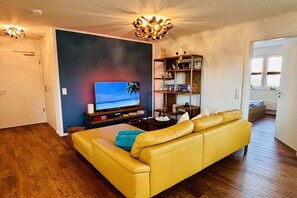 Een woonkamer met een gele bank, een televisie en een boekenkast.