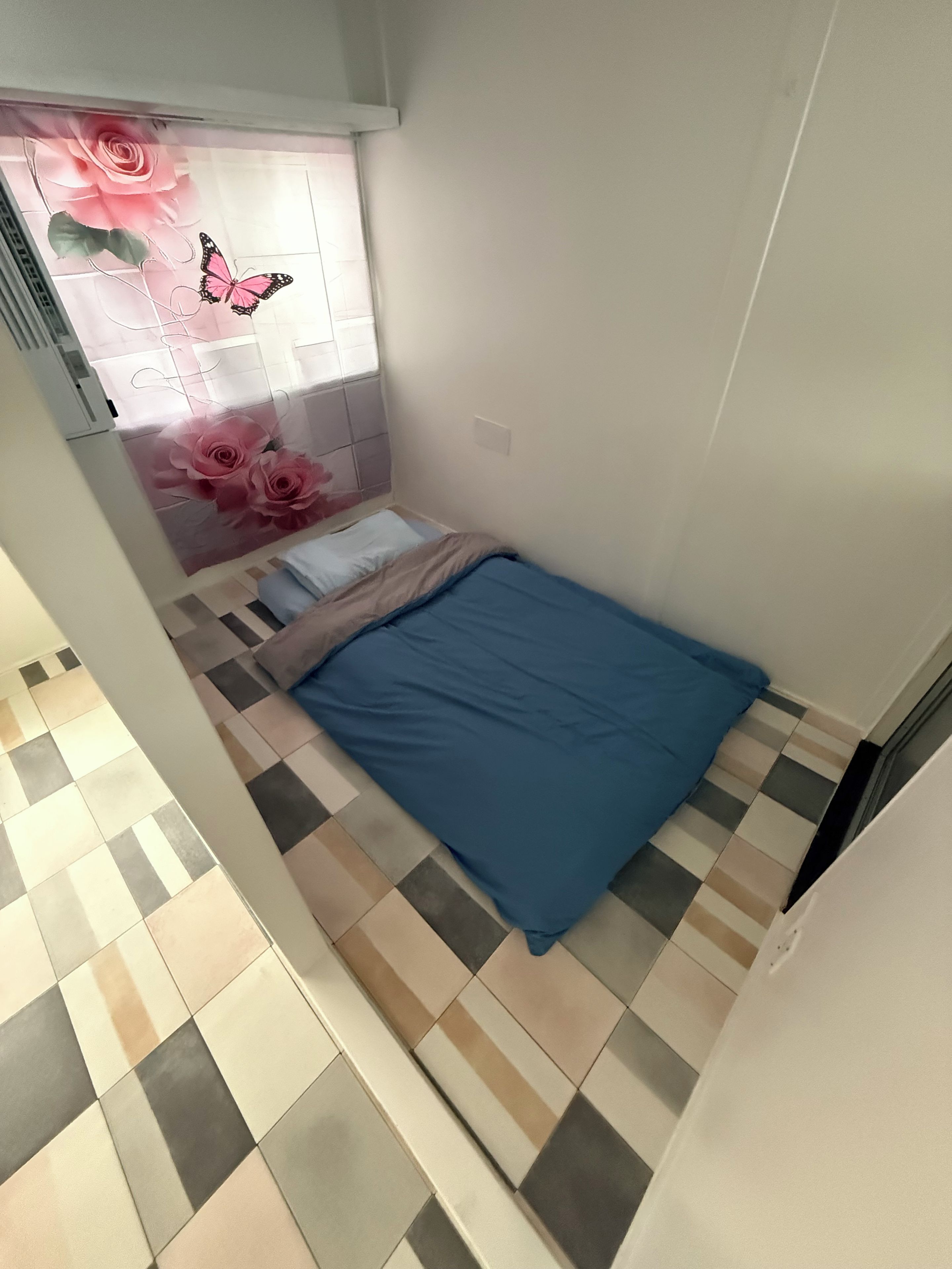 A small, single-bed room with a checkered floor, a window with a floral curtain, and a wall-mounted air conditioning unit.