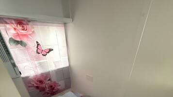A small, single-bed room with a checkered floor, a window with a floral curtain, and a wall-mounted air conditioning unit.